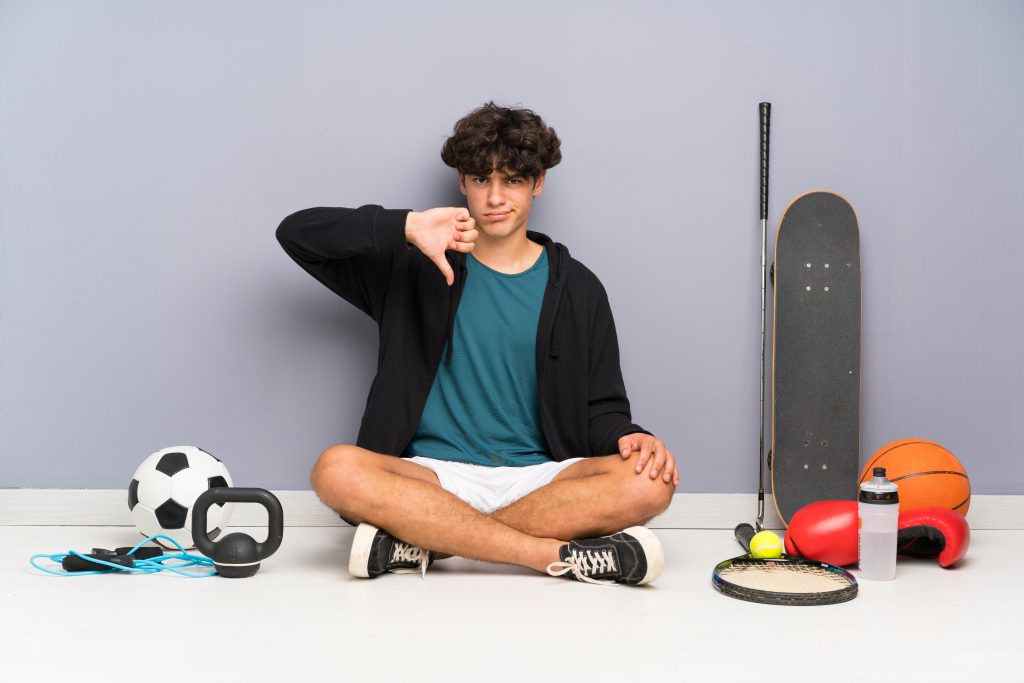 Young Sport Man Sitting On The Floor Around Many Sport Elements Showing Thumb Down Sign
